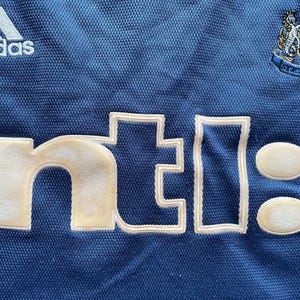 Op de afbeelding: Close-up van een blauw shirt met witte geborduurde tekst en logo's. Het Adidas-logo staat linksboven en een teamembleem rechtsboven. De tekst "nti:" staat prominent in het midden.