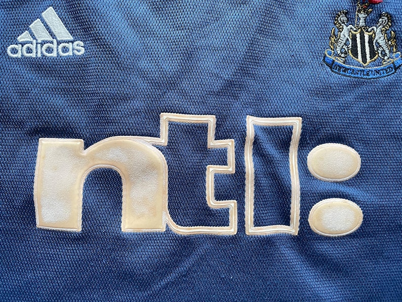 Op de afbeelding: Close-up van een blauw shirt met witte geborduurde tekst en logo's. Het Adidas-logo staat linksboven en een teamembleem rechtsboven. De tekst "nti:" staat prominent in het midden.