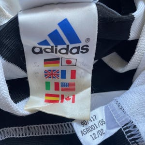 May include: White and black striped athletic jersey with a white tag that says "adidas" in blue letters. The tag also has a size chart with flags representing different countries. The tag also has the numbers "907417 ASRO001/05 12/02" printed on it.