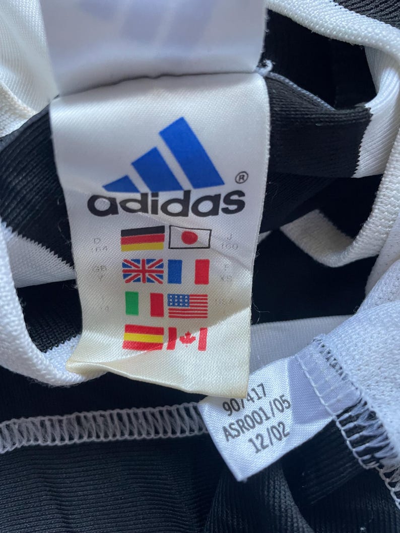 May include: White and black striped athletic jersey with a white tag that says "adidas" in blue letters. The tag also has a size chart with flags representing different countries. The tag also has the numbers "907417 ASRO001/05 12/02" printed on it.
