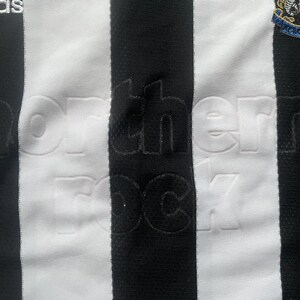 Puede incluir: Primer plano de una camiseta de f&uacute;tbol a rayas blancas y negras. La camiseta tiene la inscripci&oacute;n en relieve "northern rock". El logotipo de Adidas est&aacute; en la parte superior izquierda y el escudo del equipo en la parte superior derecha.