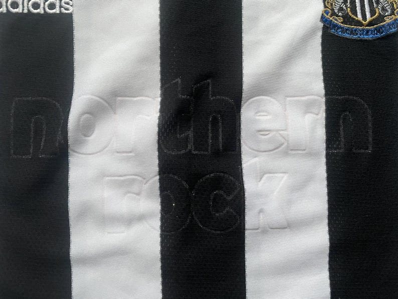 Puede incluir: Primer plano de una camiseta de f&uacute;tbol a rayas blancas y negras. La camiseta tiene la inscripci&oacute;n en relieve "northern rock". El logotipo de Adidas est&aacute; en la parte superior izquierda y el escudo del equipo en la parte superior derecha.