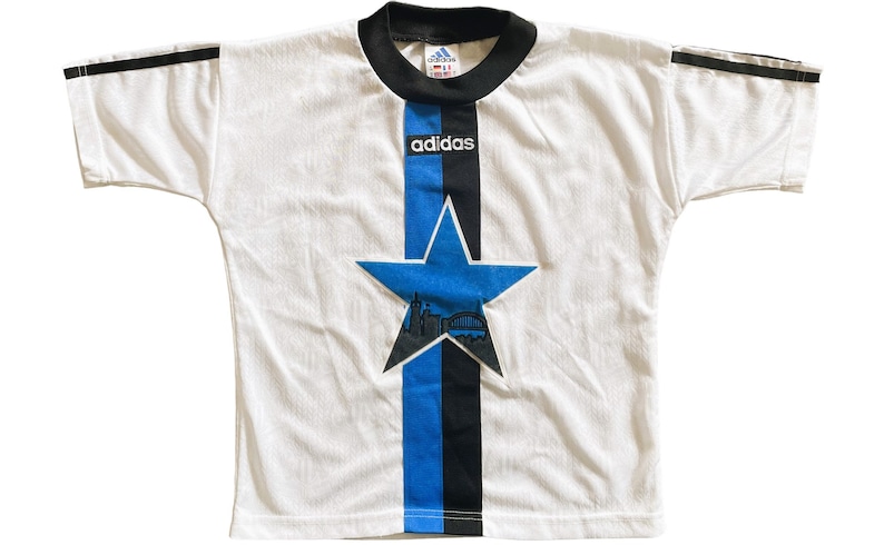 May include: White t-shirt with black and blue stripes down the front. The stripes feature a large blue star with a city skyline inside. The shirt has black adidas text and a black adidas logo on the front.