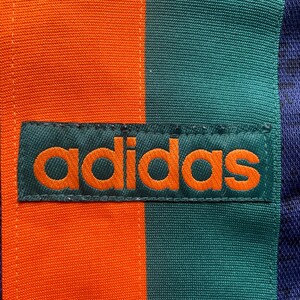 May include: Close-up of a section of fabric with the Adidas logo. The fabric is divided into vertical stripes of orange, teal, and navy blue. The Adidas logo is embroidered in orange on a teal rectangular patch.