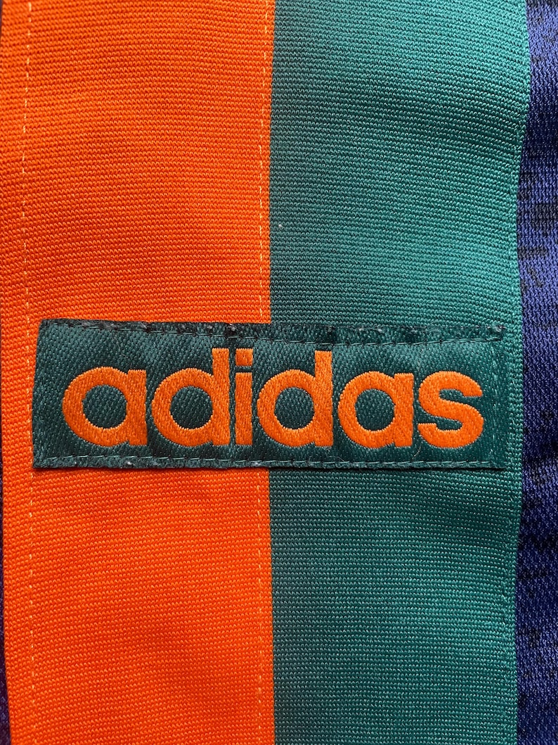 May include: Close-up of a section of fabric with the Adidas logo. The fabric is divided into vertical stripes of orange, teal, and navy blue. The Adidas logo is embroidered in orange on a teal rectangular patch.