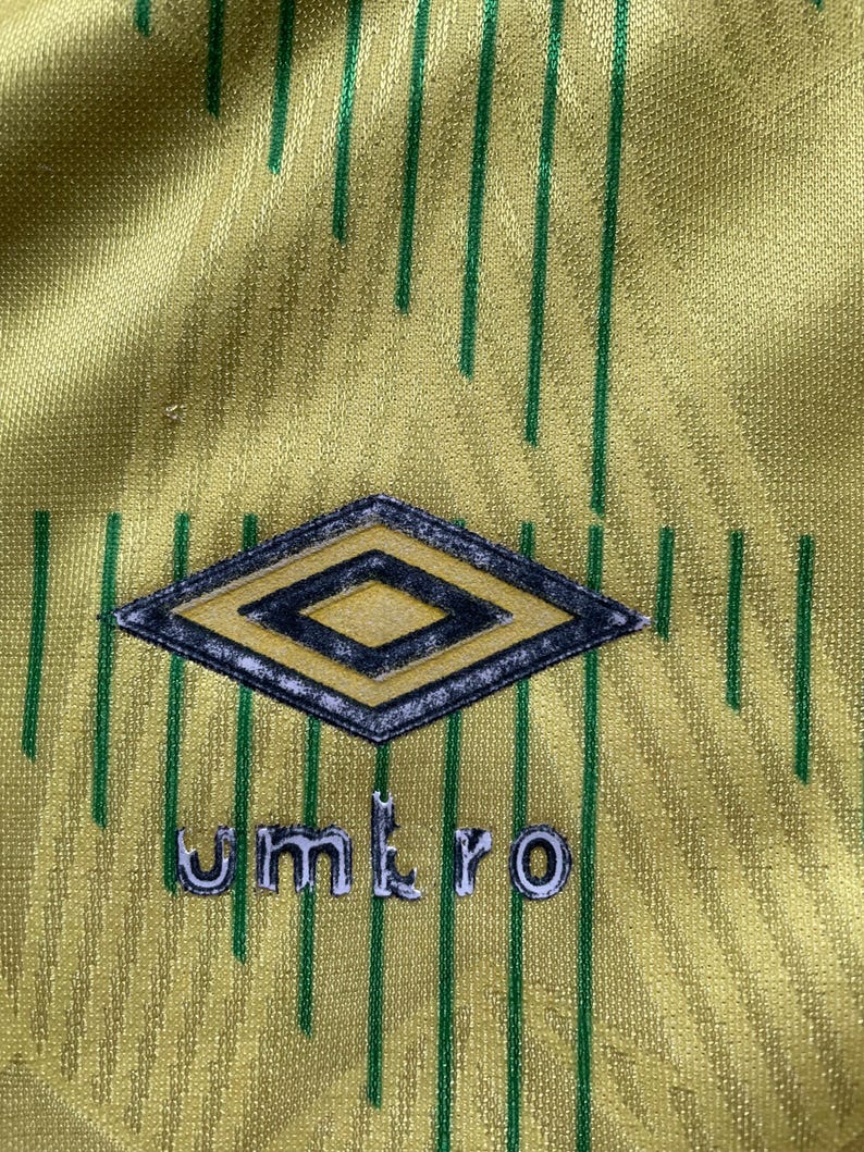May include: A yellow and green soccer jersey with a diamond-shaped logo and the word "umbro" printed on the front.