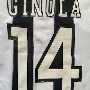 May include: A white and black striped soccer jersey with the name "GINOLA" and the number "14" printed on the back. The jersey has a classic design with bold lettering and numbering.