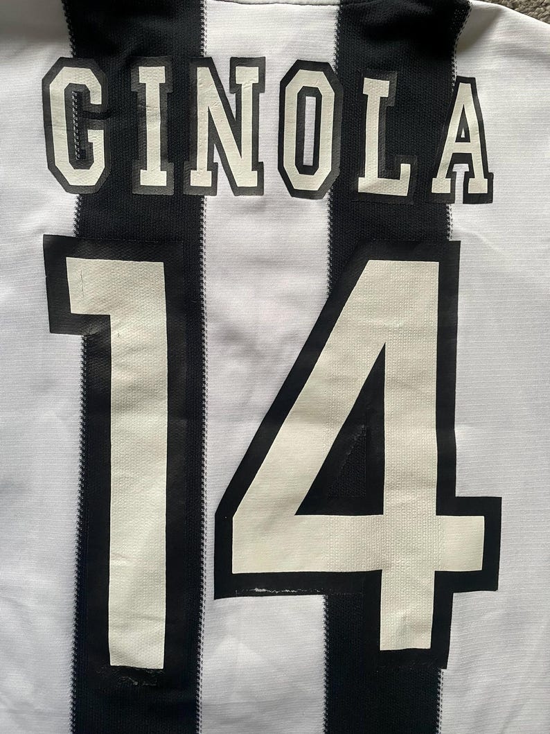 May include: A white and black striped soccer jersey with the name "GINOLA" and the number "14" printed on the back. The jersey has a classic design with bold lettering and numbering.