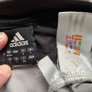 May include: Black Adidas leggings with a size tag showing sizes UK 42/44, USA 5, F 5, D 7, CDN 7, E 5, I 7, XO. The tag also says "Made in Indonesia". A white tag with the Adidas logo and flags of various countries is attached to the leggings.