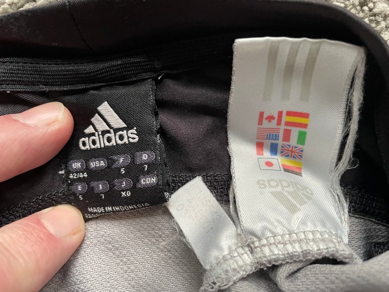 May include: Black Adidas leggings with a size tag showing sizes UK 42/44, USA 5, F 5, D 7, CDN 7, E 5, I 7, XO. The tag also says "Made in Indonesia". A white tag with the Adidas logo and flags of various countries is attached to the leggings.