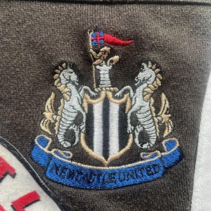May include: A brown fabric patch with an embroidered crest featuring two white horses, a black and white shield with a gold trim, and a blue banner with the text "NEWCASTLE UNITED".