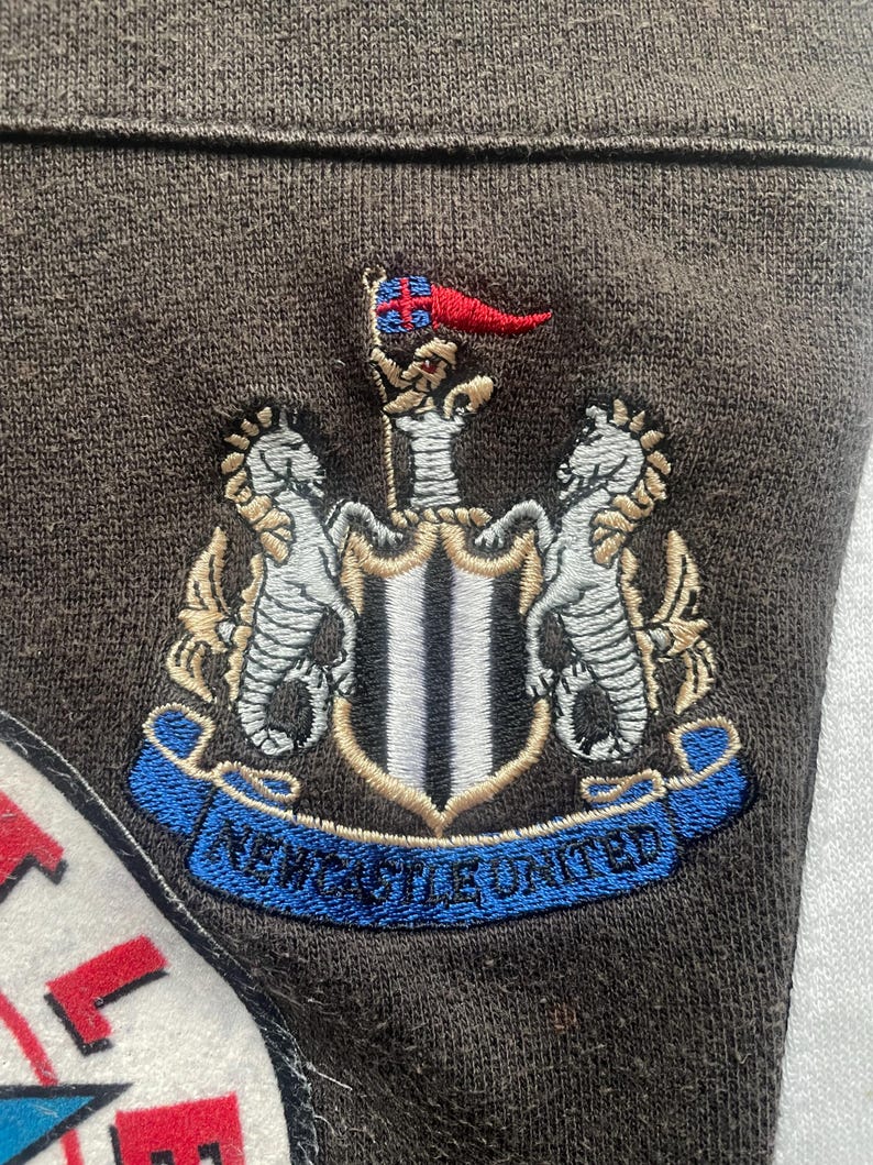 May include: A brown fabric patch with an embroidered crest featuring two white horses, a black and white shield with a gold trim, and a blue banner with the text "NEWCASTLE UNITED".