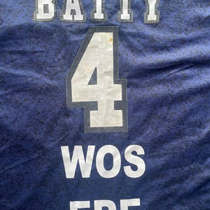 May include: Close-up of a navy blue jersey with white lettering and numbers. The word "BATTY" is at the top, with the number "4" in the center, and "WOS ERE" at the bottom. The fabric has a subtle pattern.