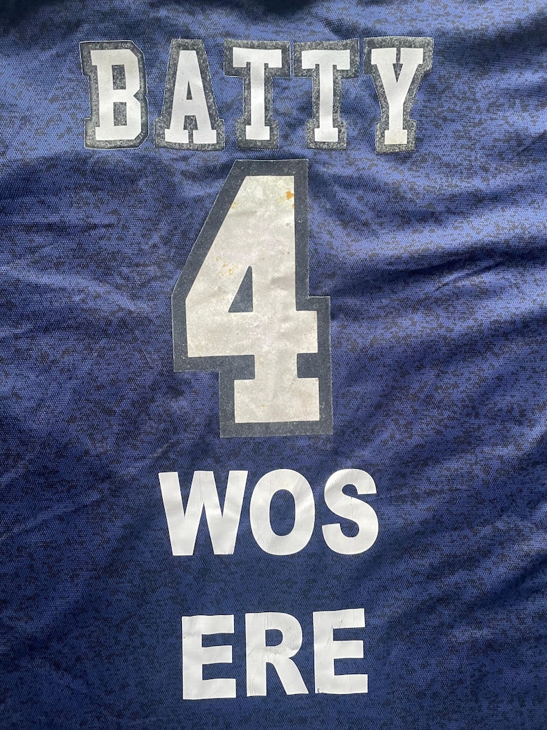 May include: Close-up of a navy blue jersey with white lettering and numbers. The word "BATTY" is at the top, with the number "4" in the center, and "WOS ERE" at the bottom. The fabric has a subtle pattern.