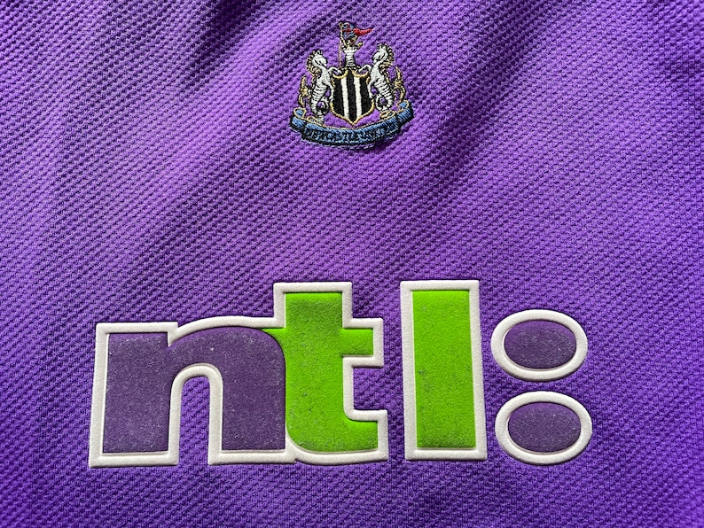 May include: A purple soccer jersey with a white and black crest and the word "ntl" in green and purple letters.