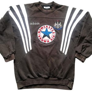 May include: A brown Adidas sweatshirt with white stripes on the sleeves and a Newcastle Brown Ale logo on the front. The logo features a star and the text "Newcastle Brown Ale".