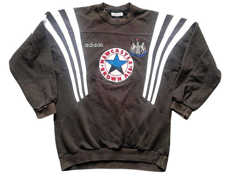 May include: A brown Adidas sweatshirt with white stripes on the sleeves and a Newcastle Brown Ale logo on the front. The logo features a star and the text "Newcastle Brown Ale".