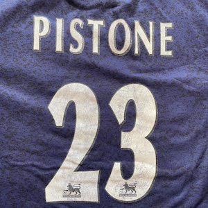 May include: A close-up of a blue jersey with the name "Pistone" and the number "23" printed in white. The jersey also features two logos of a lion with a soccer ball.