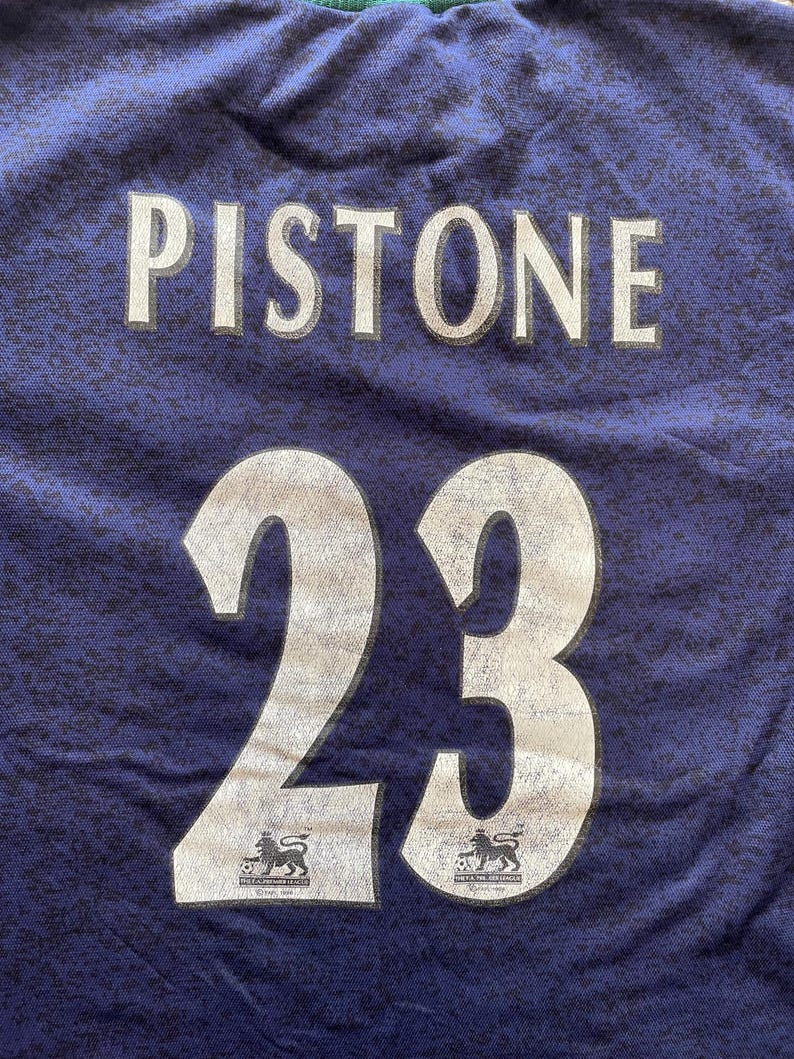May include: A close-up of a blue jersey with the name "Pistone" and the number "23" printed in white. The jersey also features two logos of a lion with a soccer ball.