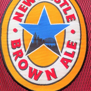 May include: A circular patch with the words "NEWCASTLE BROWN ALE" in red lettering. The patch features a blue star in the centre with a silhouette of a city skyline. The patch is on a burgundy mesh fabric.