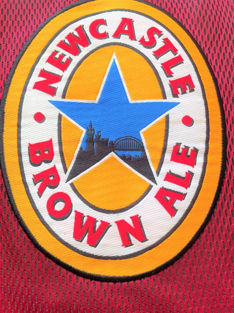 May include: A circular patch with the words "NEWCASTLE BROWN ALE" in red lettering. The patch features a blue star in the centre with a silhouette of a city skyline. The patch is on a burgundy mesh fabric.