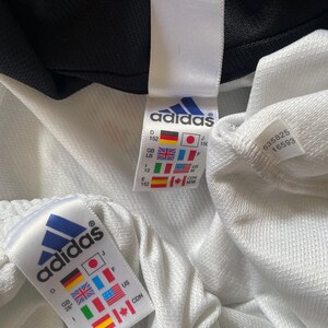 Pu&ograve; includere: Giacca da tuta Adidas bianca con cerniera nera e bordini neri. L'etichetta della taglia mostra una taglia 152 in diversi sistemi di misurazione, tra cui le taglie US, UK, EU e giapponese. L'etichetta include anche il logo Adidas e il testo "&reg;".