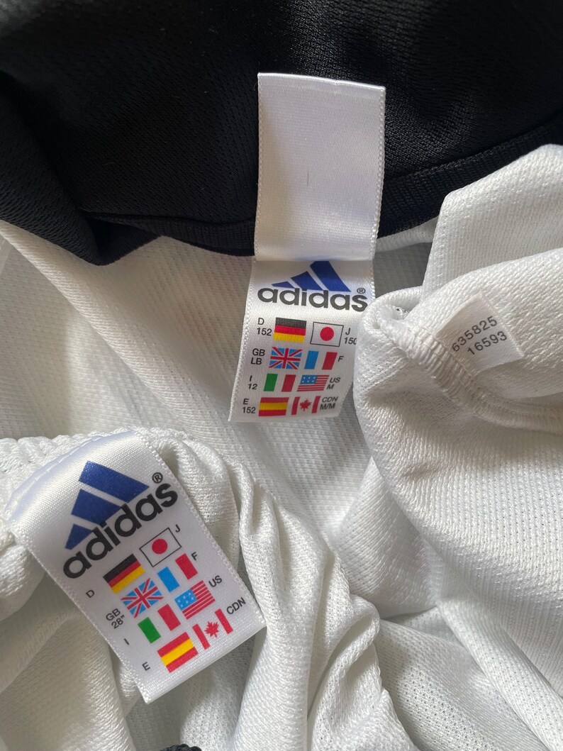 Pu&ograve; includere: Giacca da tuta Adidas bianca con cerniera nera e bordini neri. L'etichetta della taglia mostra una taglia 152 in diversi sistemi di misurazione, tra cui le taglie US, UK, EU e giapponese. L'etichetta include anche il logo Adidas e il testo "&reg;".