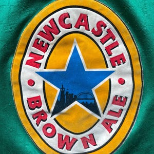 K&ouml;nnte beinhalten: Ein kreisf&ouml;rmiger Aufn&auml;her mit dem Schriftzug "NEWCASTLE BROWN ALE" in roter Schrift. Das Design umfasst einen gelben Rand, einen blauen Stern und eine Silhouette der Skyline einer Stadt und einer Br&uuml;cke.