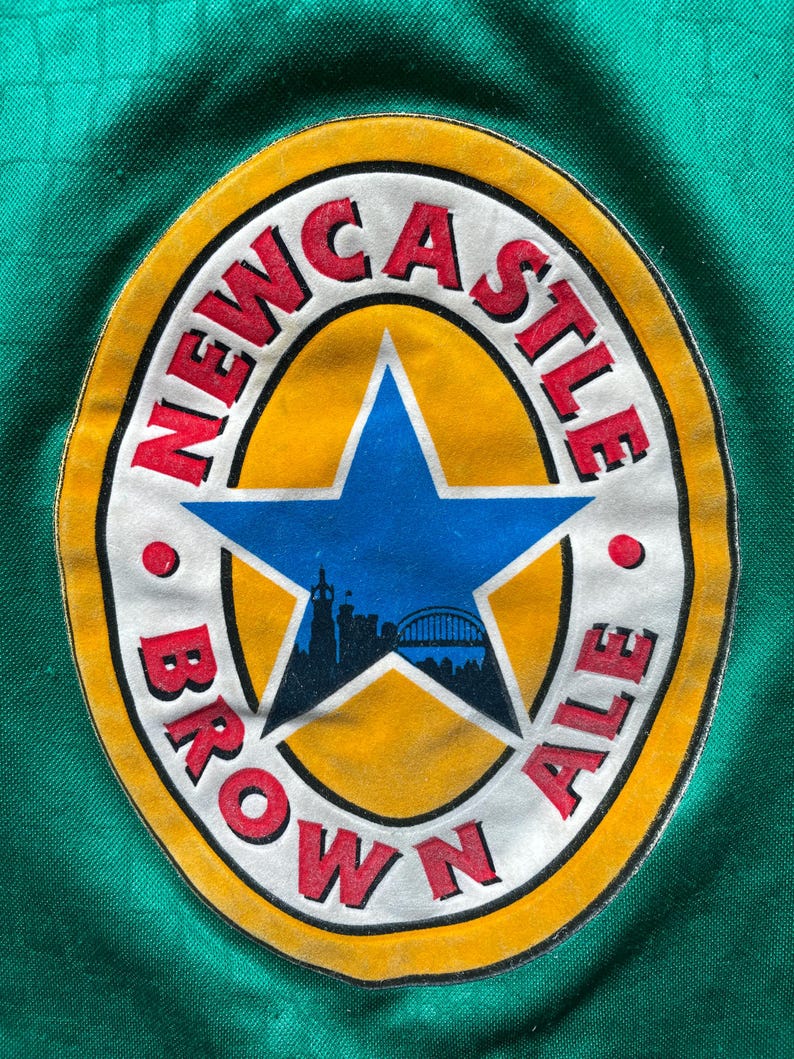 K&ouml;nnte beinhalten: Ein kreisf&ouml;rmiger Aufn&auml;her mit dem Schriftzug "NEWCASTLE BROWN ALE" in roter Schrift. Das Design umfasst einen gelben Rand, einen blauen Stern und eine Silhouette der Skyline einer Stadt und einer Br&uuml;cke.