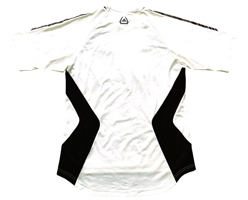Pode incluir: Camisa esportiva branca com detalhes em preto nas laterais e mangas. A camisa tem gola redonda e o logotipo da Adidas na parte superior das costas. As mangas t&ecirc;m tr&ecirc;s listras pretas.