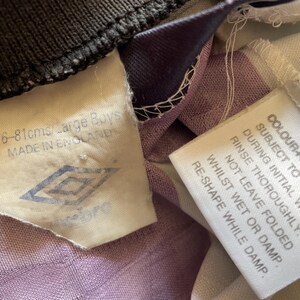 Pode incluir: Uma camisa de futebol branca e roxa com uma etiqueta branca que diz "COLOURFAST DYES ARE SUBJECT TO 'BLEEDING' DURING INITIAL WASHES. RINSE THOROUGHLY. DO NOT LEAVE FOLDED WHILST WET OR DAMP. RE-SHAPE WHILE DAMP." e uma etiqueta branca com um logotipo preto que diz "76-81cms/Large Boys MADE IN ENGLAND".