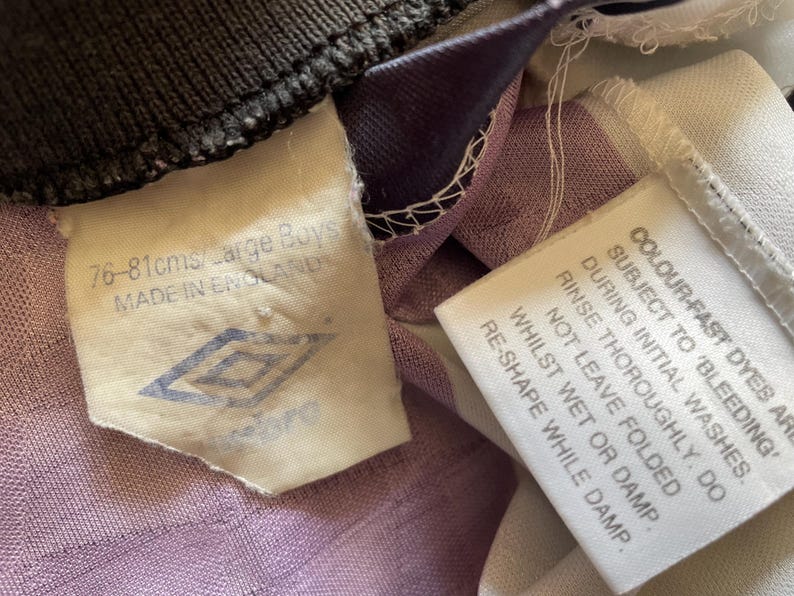 Pode incluir: Uma camisa de futebol branca e roxa com uma etiqueta branca que diz "COLOURFAST DYES ARE SUBJECT TO 'BLEEDING' DURING INITIAL WASHES. RINSE THOROUGHLY. DO NOT LEAVE FOLDED WHILST WET OR DAMP. RE-SHAPE WHILE DAMP." e uma etiqueta branca com um logotipo preto que diz "76-81cms/Large Boys MADE IN ENGLAND".