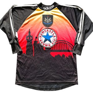 May include: Black and red Newcastle United soccer jersey with white stripes on the sleeves. The jersey features the Adidas logo, a crest, and a cityscape design with the words "Newcastle Brown Ale."