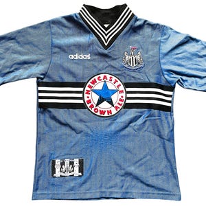 May include: Light blue Newcastle United football shirt with black and white detailing. Includes the Adidas logo, a Newcastle Brown Ale emblem, and the team crest. Short-sleeved with a V-neck collar.