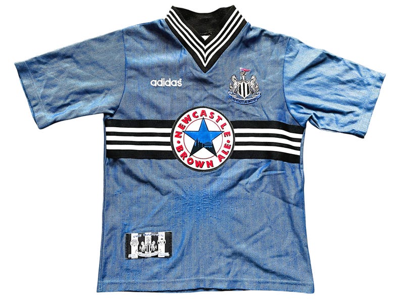 May include: Light blue Newcastle United football shirt with black and white detailing. Includes the Adidas logo, a Newcastle Brown Ale emblem, and the team crest. Short-sleeved with a V-neck collar.