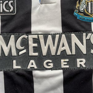K&ouml;nnte beinhalten: Nahaufnahme eines schwarz-wei&szlig; gestreiften Fu&szlig;balltrikots. Das Trikot zeigt den Text "McEwan's Lager" in Wei&szlig;. Das Wappen von Newcastle United ist sichtbar, ebenso wie das Asics-Logo. Ein klassisches Design.