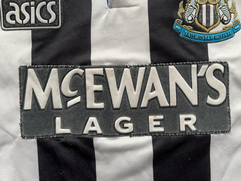 K&ouml;nnte beinhalten: Nahaufnahme eines schwarz-wei&szlig; gestreiften Fu&szlig;balltrikots. Das Trikot zeigt den Text "McEwan's Lager" in Wei&szlig;. Das Wappen von Newcastle United ist sichtbar, ebenso wie das Asics-Logo. Ein klassisches Design.