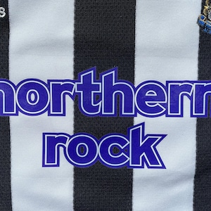 Pode incluir: Uma camisa de futebol listrada branca e preta com um logotipo "northern rock" azul e branco na frente. A camisa tamb&eacute;m tem um logotipo da Adidas no lado esquerdo e um bras&atilde;o de equipe no lado direito.