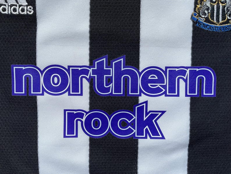 Pode incluir: Uma camisa de futebol listrada branca e preta com um logotipo "northern rock" azul e branco na frente. A camisa tamb&eacute;m tem um logotipo da Adidas no lado esquerdo e um bras&atilde;o de equipe no lado direito.