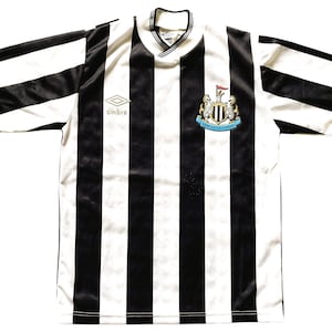 Op de afbeelding: Een zwart-wit gestreept voetbalshirt met korte mouwen. Het shirt heeft het Newcastle United-embleem en het Umbro-logo. De kraag is een V-hals.