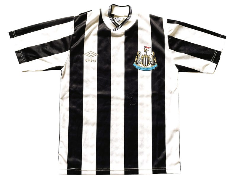 Op de afbeelding: Een zwart-wit gestreept voetbalshirt met korte mouwen. Het shirt heeft het Newcastle United-embleem en het Umbro-logo. De kraag is een V-hals.