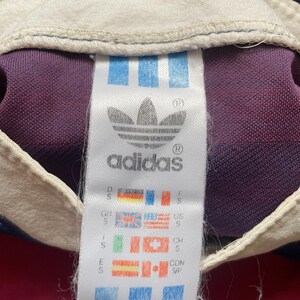 May include: An Adidas label featuring the trefoil logo and text, with flags representing different countries. The label is white with blue stripes, set against a burgundy, navy, and white fabric background.