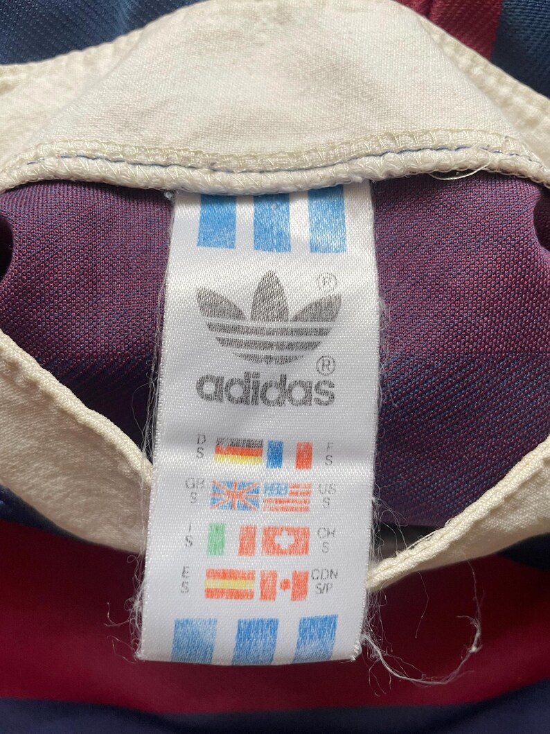 May include: An Adidas label featuring the trefoil logo and text, with flags representing different countries. The label is white with blue stripes, set against a burgundy, navy, and white fabric background.