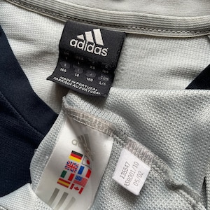 Puede incluir: Una camiseta gris Adidas con un logotipo blanco y negro y una etiqueta de talla. La etiqueta muestra las tallas en Reino Unido, EE. UU., UE y CON. La etiqueta tambi&eacute;n dice "Hecho en Portugal" y "Fabrique au Portugal".