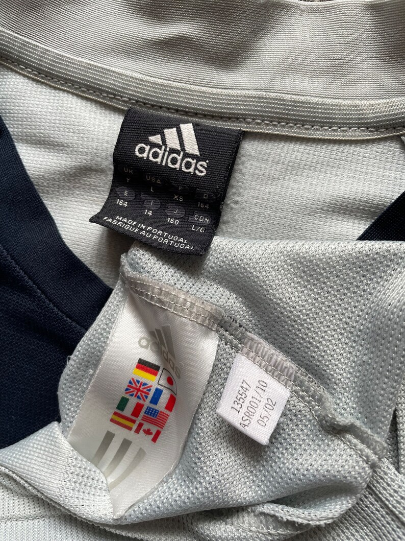 Puede incluir: Una camiseta gris Adidas con un logotipo blanco y negro y una etiqueta de talla. La etiqueta muestra las tallas en Reino Unido, EE. UU., UE y CON. La etiqueta tambi&eacute;n dice "Hecho en Portugal" y "Fabrique au Portugal".