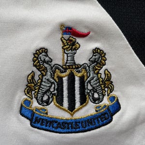Pode incluir: Bras&atilde;o bordado do Newcastle United em tecido branco e preto. O bras&atilde;o apresenta um escudo com listras pretas e brancas, dois cavalos-marinhos e uma faixa com "NEWCASTLE UNITED". Uma pequena bandeira com detalhes vermelhos e azuis no topo.