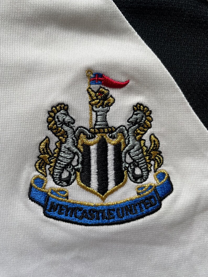 Pode incluir: Bras&atilde;o bordado do Newcastle United em tecido branco e preto. O bras&atilde;o apresenta um escudo com listras pretas e brancas, dois cavalos-marinhos e uma faixa com "NEWCASTLE UNITED". Uma pequena bandeira com detalhes vermelhos e azuis no topo.