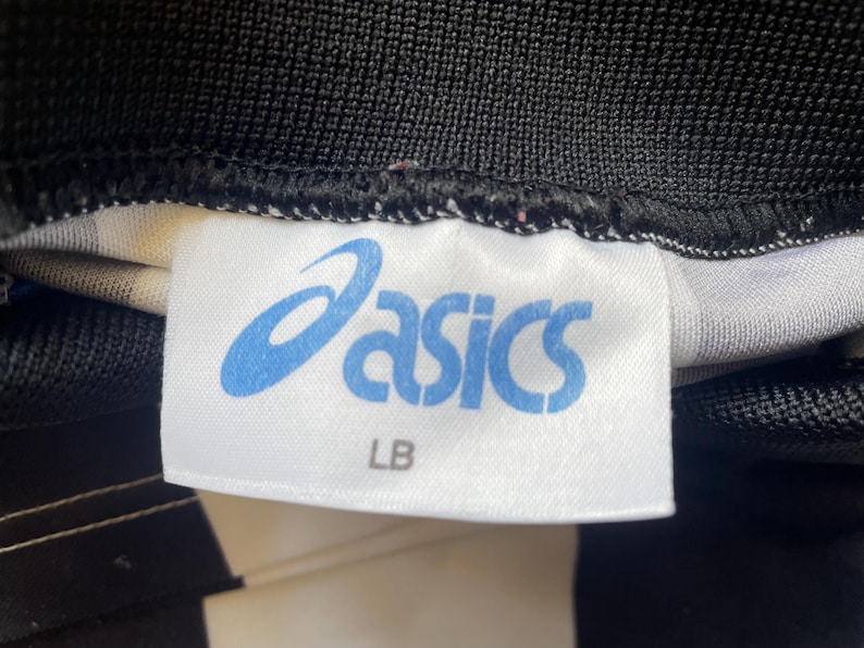 May include: Close-up of a white tag with the blue "asics" logo and the letters "LB". The tag is sewn onto black and white fabric.