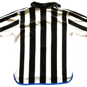 May include: A black and white striped referee shirt with a black collar and blue trim. The short-sleeved shirt has a mesh-like texture and is viewed from the back. The stripes are vertical and evenly spaced.
