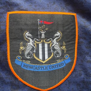 May include: Embroidered patch featuring the Newcastle United crest. The crest is shield-shaped with a black and white striped center, flanked by two seahorses. The patch has an orange border and a blue banner with the text "NEWCASTLE UNITED".